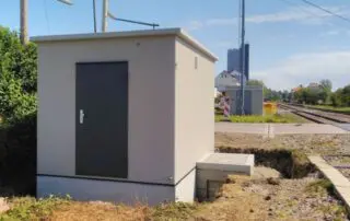 Ein kleines, graues Versorgungsgebäude mit einer dunklen Tür steht neben einem Bahngleis. Der Boden daneben ist ausgehoben, und Betonfertigteile bilden eine Betonplattform, die sich vom Gebäude aus über den offenen Graben erstreckt.