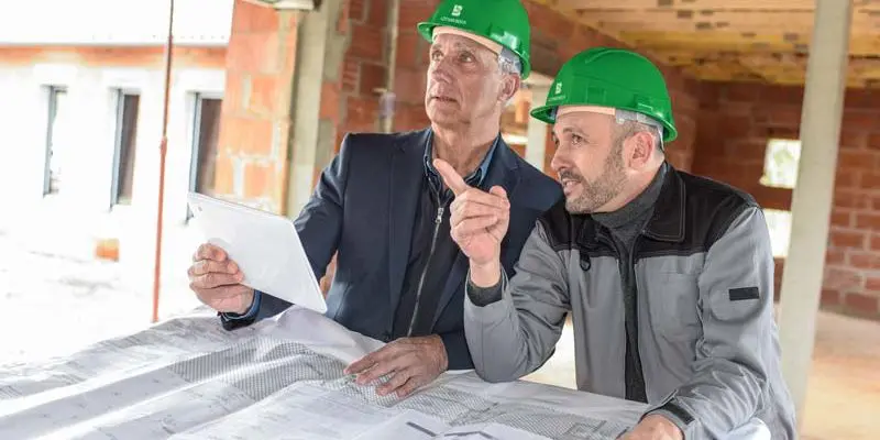 Zwei Männer mit grünen Schutzhelmen prüfen Baupläne auf einer Baustelle. Ein Mann hält ein Klemmbrett in der Hand, während der andere in Richtung der Pläne gestikuliert. Im Hintergrund sind unfertige Ziegelwände und Holzbalken zu sehen.